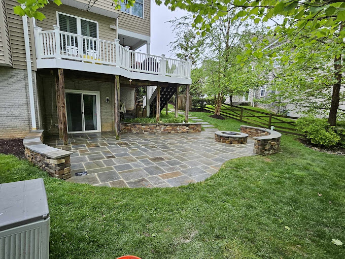 Beautiful stone patio built by ProlanscapesMD with a built-in firepit and retaining wall, nestled beneath a raised deck in a lush Maryland backyard, perfect for outdoor gatherings.